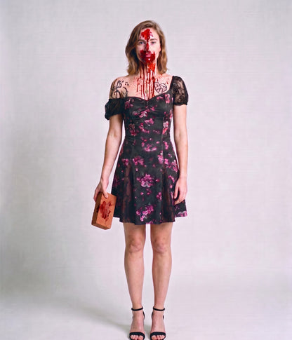 Woman wearing a dark floral off-shoulder mini dress with fake blood Halloween makeup and black marker tattoos, holding a blood-smeared brick, full-length front view on a grey background