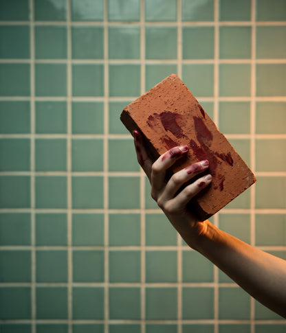 Hand holding a brick smeared with fake blood in front of a green tiled wall